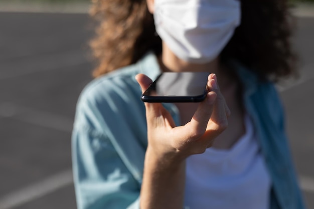 Foto mujer caucásica en las calles de la ciudad durante el día, usando una máscara facial contra el coronavirus covid19 sosteniendo y hablando en su teléfono inteligente, concéntrese en primer plano