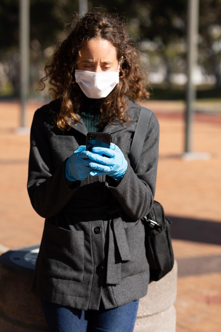 Foto mujer caucásica en las calles de la ciudad durante el día, usando una máscara facial y guantes sanitarios contra el coronavirus covid19, de pie y usando su teléfono inteligente