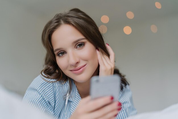 Mujer caucásica en la cama usando un teléfono inteligente moderno con auriculares para ver videos y escuchar audio en redes sociales Concepto de tecnología y entretenimiento