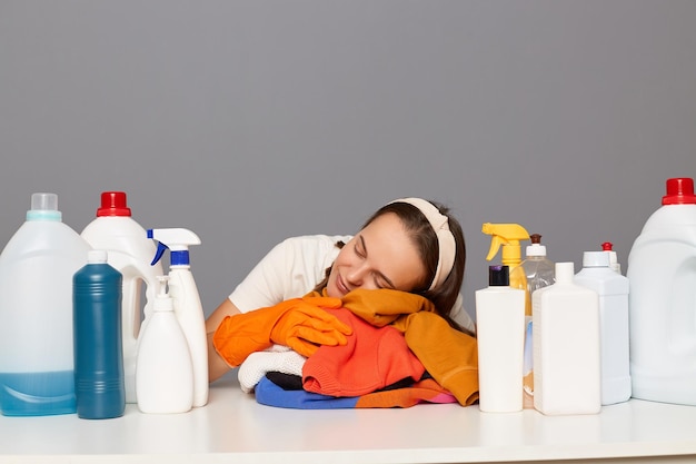 Una mujer caucásica cansada y soñolienta usa guantes protectores de goma y una banda para la cabeza rodeada de botellas de detergentes para lavar la ropa apoyada en la mesa para dormir se siente cansada y exhausta