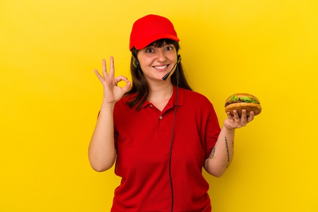 Mujer caucásica con curvas joven que sostiene la hamburguesa aislada en el fondo amarillo alegre y confiado que muestra el gesto aceptable.