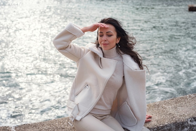 Una mujer caucásica despreocupada en ropa beige que disfruta de la vista del mar en un día caluroso y ventoso.