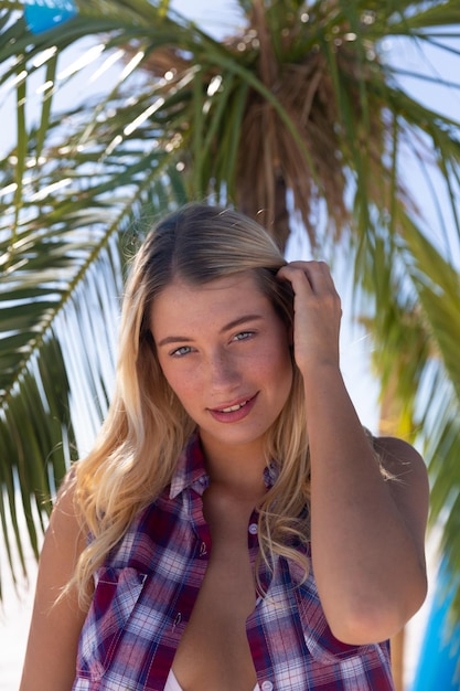 Mujer caucásica disfrutando del tiempo en la playa, de pie junto a una palmera, mirando a la cámara y sonriendo