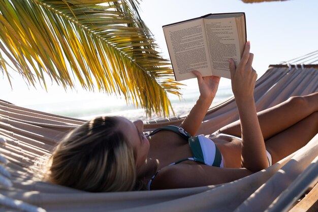 Mujer caucásica disfrutando del tiempo en la playa, recostada en una hamaca y leyendo un libro