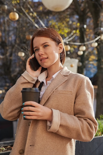 Mujer caucásica en elegante abrigo beige llamando por teléfono inteligente tomando café