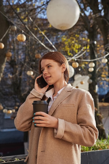 Mujer caucásica en elegante abrigo beige llamando por teléfono inteligente tomando café