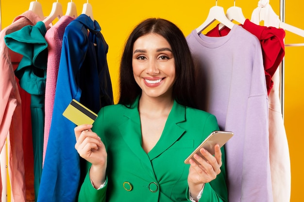 Mujer caucásica emocionada usando teléfono celular y tarjeta de crédito para ir de compras posando cerca del perchero y