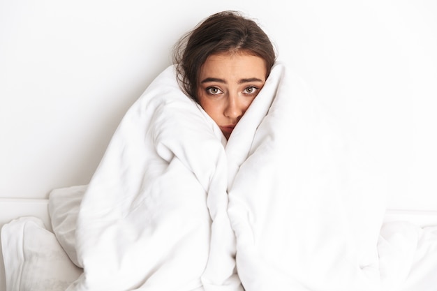 Mujer caucásica envuelta en una manta sonriendo y sentada en la cama en el apartamento