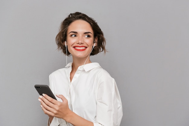 Mujer caucásica escuchando música en el teléfono inteligente a través de auriculares, aislado sobre una pared gris