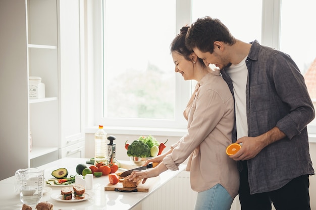 Mujer caucásica está cortando pan para sándwiches mientras su amante la abraza y sonríe