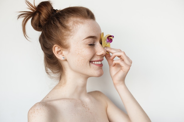 Mujer caucásica está cubriendo su ojo con una flor posando con los hombros desnudos en una pared blanca de estudio