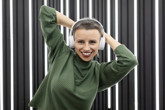 Mujer caucásica con estilo en auriculares con una amplia sonrisa en el fondo de una pared retroiluminada