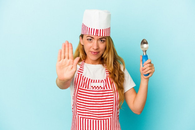 Mujer caucásica del fabricante del helado que sostiene una bola del helado aislada en el fondo azul que se coloca con la mano extendida que muestra la señal de pare, que le impide.