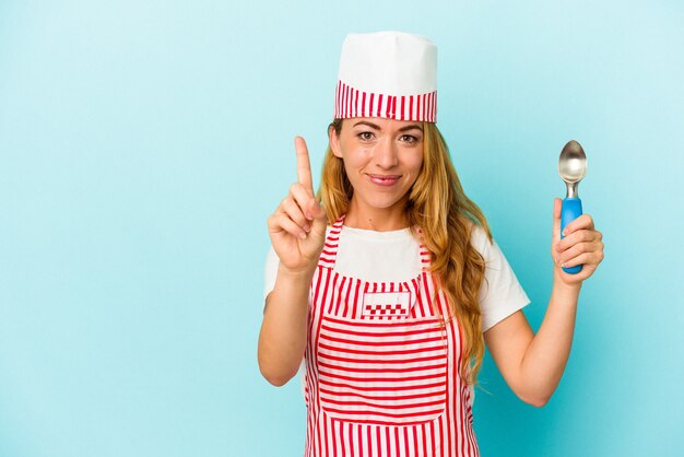 Mujer caucásica del fabricante de helado que sostiene una bola del helado aislada en fondo azul que muestra el número uno con el dedo.