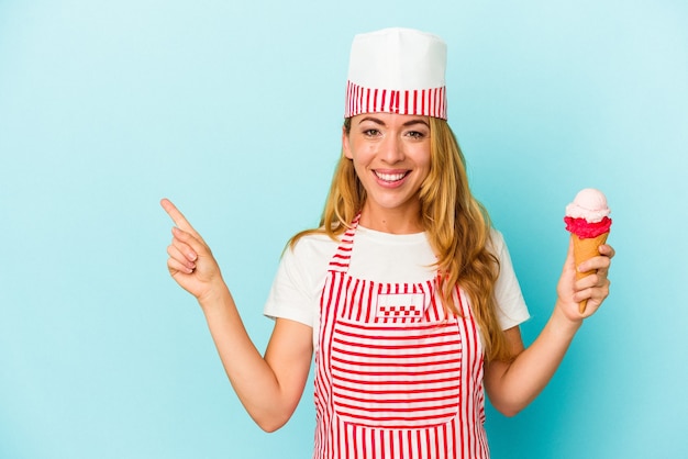 Mujer caucásica fabricante de helado sosteniendo un helado aislado sobre fondo azul sonriendo y apuntando a un lado, mostrando algo en el espacio en blanco.