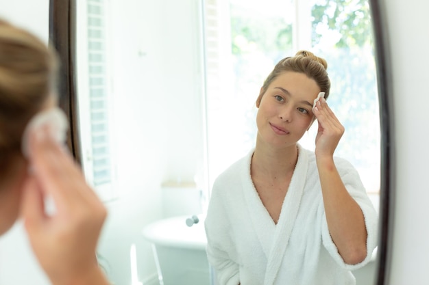 Mujer caucásica feliz en bata de baño limpiando la cara con almohadilla de algodón en el baño soleado