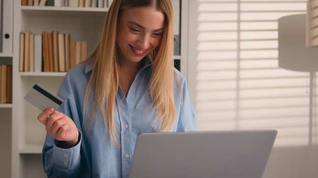 Mujer caucásica feliz y sonriente paga compras en línea con portátil y tarjeta de crédito bancaria satisfecha