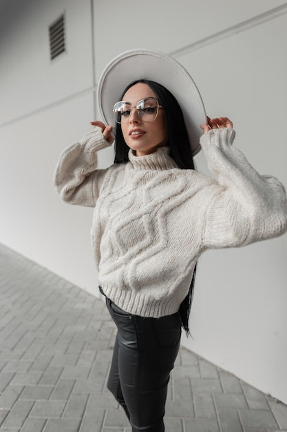 Mujer caucásica feliz con sonrisa y anteojos antiguos en un moderno suéter de punto beige y sombrero camina por la ciudad