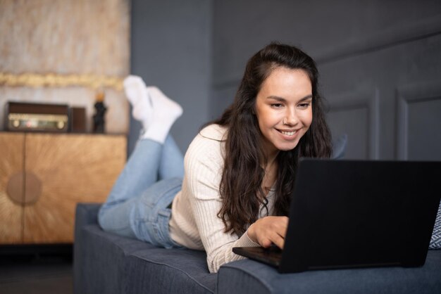 Foto mujer caucásica feliz usando computadora portátil trabajando o estudiando en línea acostado en el sofá en casa espacio libre