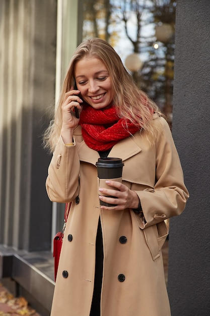 Mujer caucásica en gabardina beige llamando por teléfono inteligente con taza de café