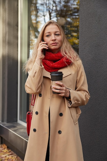 Mujer caucásica en gabardina beige llamando por teléfono inteligente con taza de café