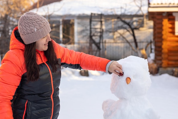 Mujer caucásica haciendo ojos y nariz de muñeco de nieve durante el día mientras camina en el patio trasero con casas y luz solar brillante en el fondo Los padres pasan tiempo con los niños