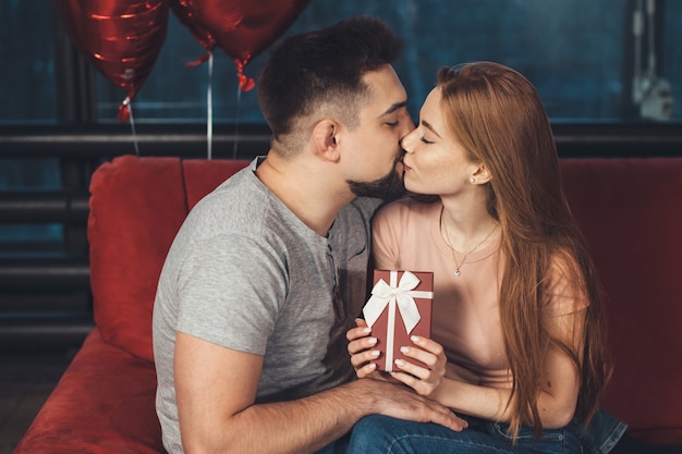 Mujer caucásica de jengibre está besando a su amante después de recibir un regalo en el día de San Valentín posando en un sofá rojo
