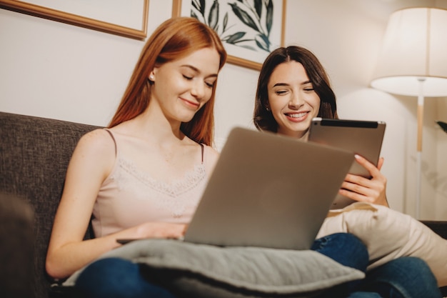 La mujer caucásica de jengibre con pecas está usando una computadora portátil cerca de su amiga morena mientras está sentada en el sofá