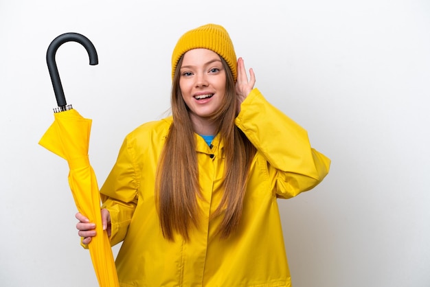 Mujer caucásica joven con abrigo impermeable y paraguas aislado en fondo blanco escuchando algo poniendo la mano en la oreja