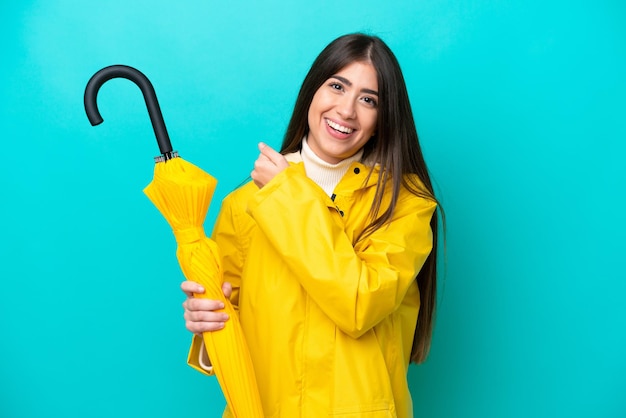 Mujer caucásica joven con abrigo impermeable y paraguas aislado sobre fondo azul celebrando una victoria