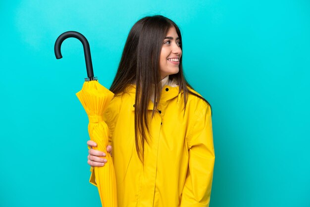 Mujer caucásica joven con abrigo impermeable y paraguas aislado sobre fondo azul mirando hacia el lado y sonriendo