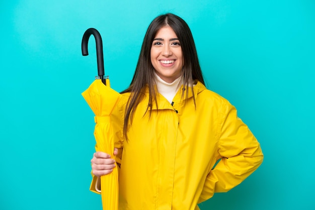 Mujer caucásica joven con abrigo impermeable y paraguas aislado sobre fondo azul posando con los brazos en la cadera y sonriendo