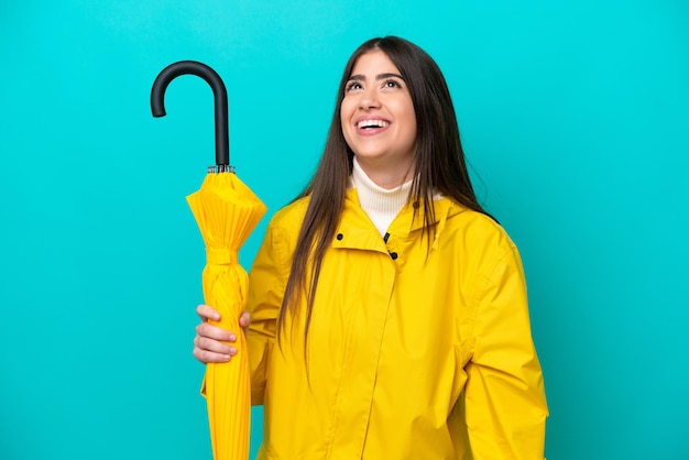 Mujer caucásica joven con abrigo impermeable y paraguas aislado sobre fondo azul riendo