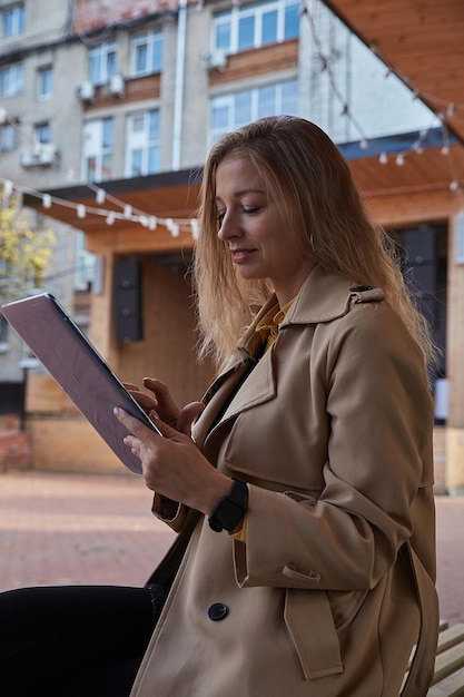 Mujer caucásica joven en abrigo usando tableta al aire libre en un día soleado, leyendo libros electrónicos, viendo películas. atractivo retrato callejero femenino en otoño o primavera. estilo de vida otoñal, tecnología de comunicación moderna.