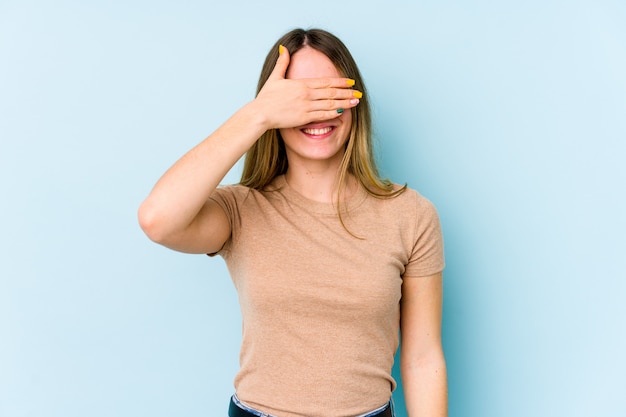 Foto la mujer caucásica joven aislada en azul cubre ojos con las manos, sonríe ampliamente esperando una sorpresa.