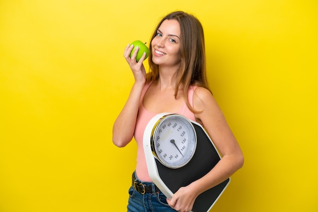 Mujer caucásica joven aislada de fondo amarillo con máquina de pesaje y con una manzana