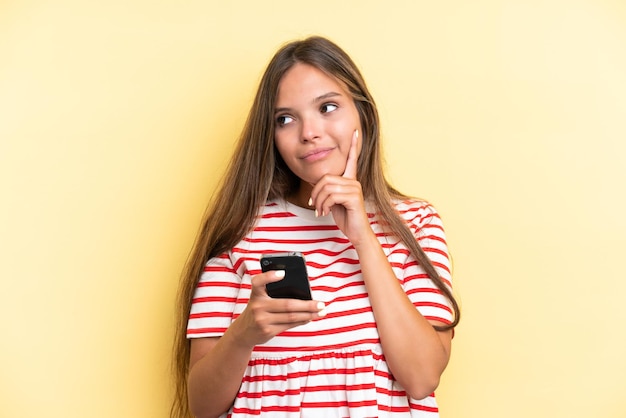 Mujer caucásica joven aislada de fondo amarillo usando teléfono móvil y pensando