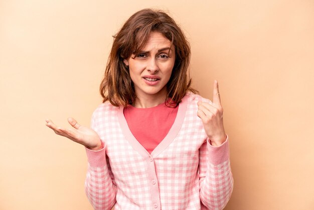 Mujer caucásica joven aislada de fondo beige sosteniendo y mostrando un producto a mano