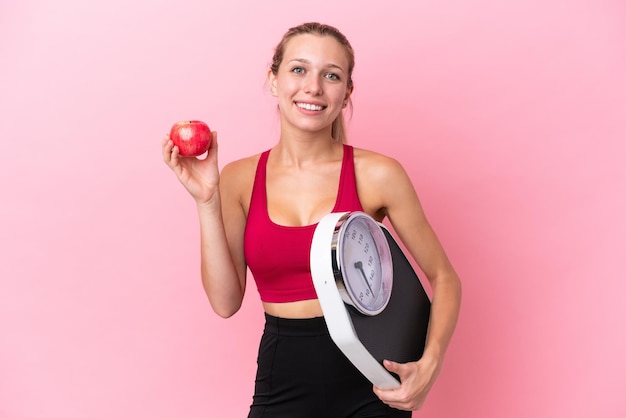 Mujer caucásica joven aislada de fondo rosa con máquina de pesaje y con una manzana