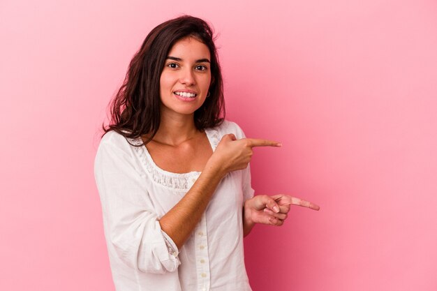 Mujer caucásica joven aislada en un fondo rosado sorprendida señalando con los dedos índices a un espacio de copia.