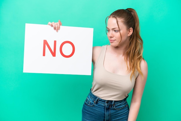 Mujer caucásica joven aislada de fondo verde sosteniendo un cartel con el texto NO