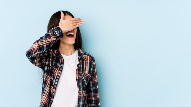 La mujer caucásica joven aislada en la pared azul cubre ojos con las manos, sonríe ampliamente esperando una sorpresa.