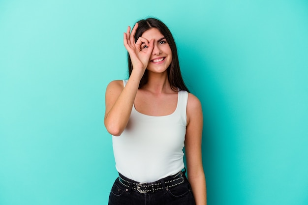 Mujer caucásica joven aislada en la pared azul emocionada manteniendo el gesto ok en el ojo.