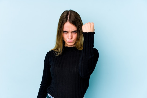 Mujer caucásica joven aislada en la pared azul que muestra el puño, la expresión facial agresiva.