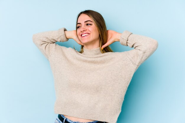 Mujer caucásica joven aislada en la pared azul que se siente confiada, con las manos detrás de la cabeza.