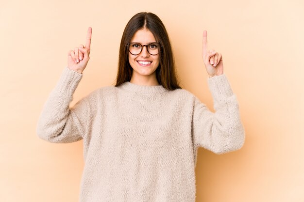 La mujer caucásica joven aislada en la pared beige indica con ambos dedos delanteros para arriba que muestran un espacio en blanco.