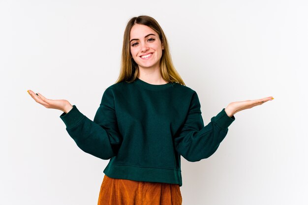 La mujer caucásica joven aislada en la pared blanca hace escala con los brazos, se siente feliz y confiada.