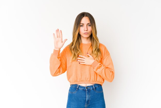 Mujer caucásica joven aislada en la pared blanca que hace un juramento, poniendo la mano en el pecho.