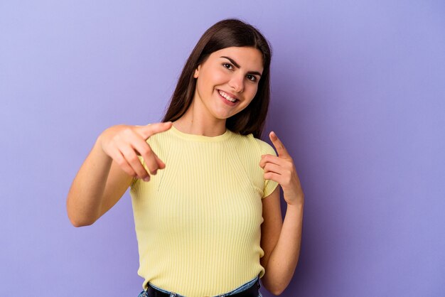 Mujer caucásica joven aislada en la pared púrpura sonrisas alegres apuntando al frente.