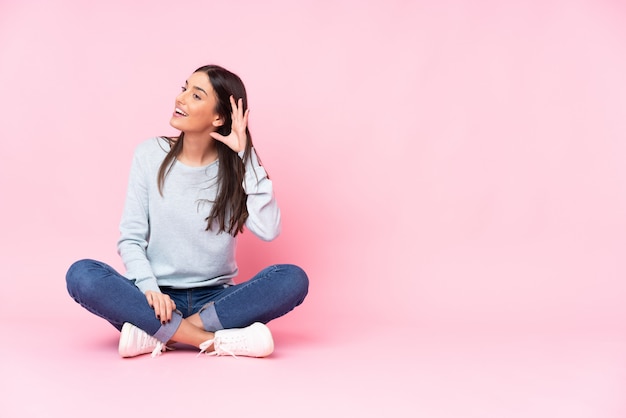 Mujer caucásica joven aislada en la pared rosa escuchando algo poniendo la mano en la oreja